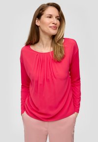 Langärmliges Oberteil in leuchtendem Pink, mit Rundhalsausschnitt und gefältetem Vorderdesign. Kombiniert mit hellpinkfarbigen Hosen. Glatte Stofftextur.