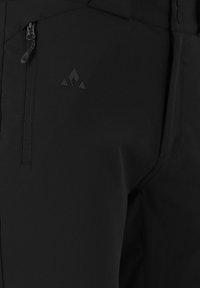 Musta pehmeäkuorihousut, joissa on vetoketjullinen sivutasku, tekstuuri pintakäsittely ja hillitty logo kankaassa, suunniteltu ulkoilmaaktiviteetteihin.