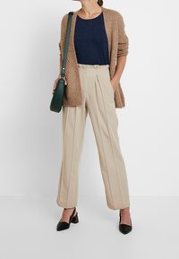 Pantaloni alti beige con pieghe laterali, abbinati a una blusa blu marina e a un cardigan di lana grosso marrone. Scarpe nere con tacco a blocco. Borsa verde.