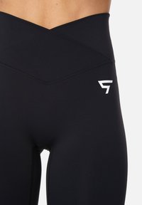 Zwarte high-waisted leggings met een crisscross taillebandontwerp, voorzien van een wit logo op de linker dij en een gladde textuur.