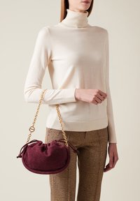 Borsa a tracolla di colore bordeaux, con texture, chiusura a coulisse e forma arrotondata, tenuta in mano accanto a un maglione chiaro e pantaloni a fantasia.
