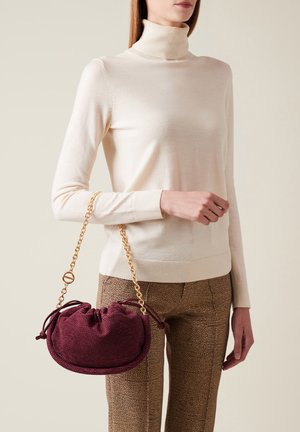 Sac à main texturé bourgogne avec une chaîne, fermeture à cordon, et forme arrondie, tenu à la main à côté d'un pull léger et d'un pantalon à motifs.