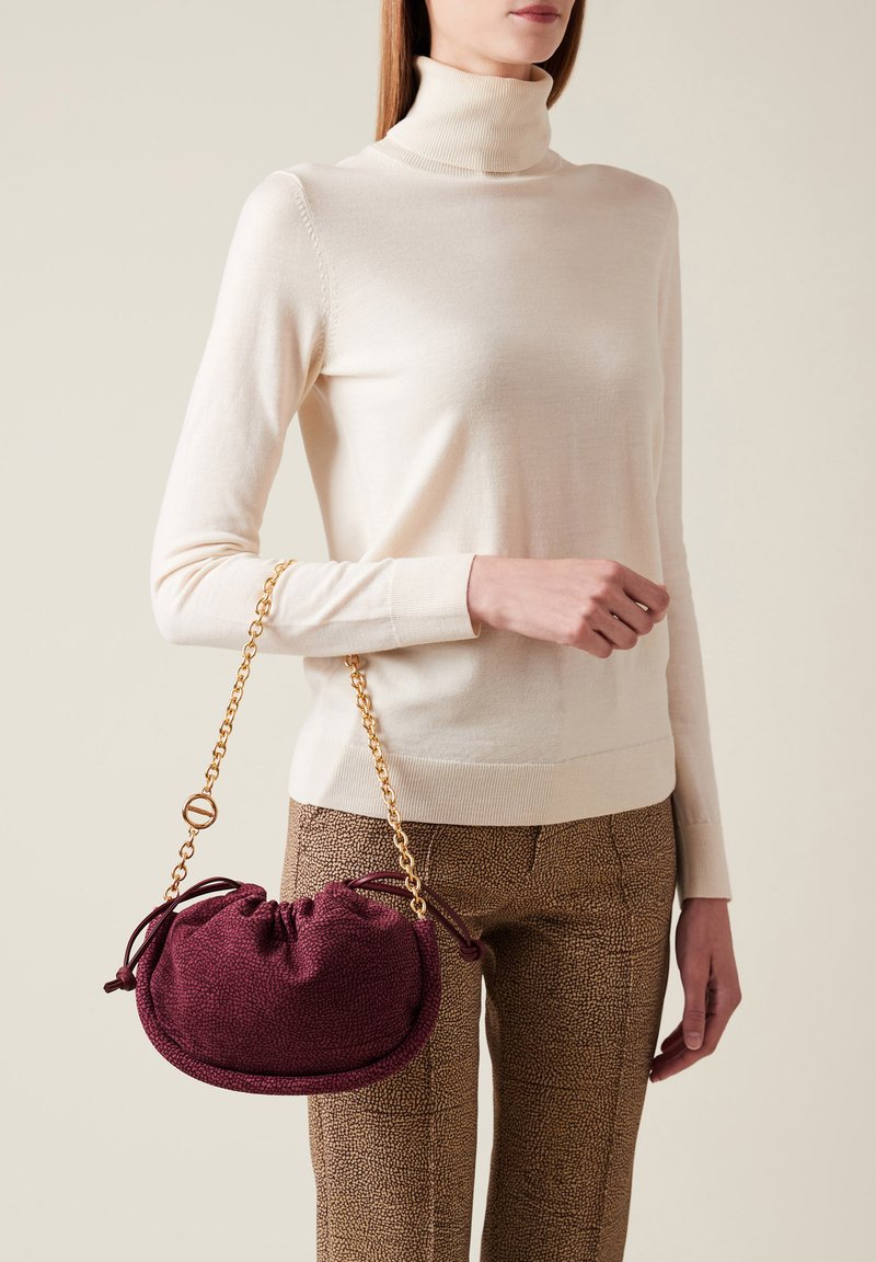 Borsa a tracolla di colore bordeaux, con texture, chiusura a coulisse e forma arrotondata, tenuta in mano accanto a un maglione chiaro e pantaloni a fantasia.