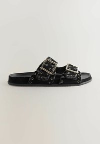 Sorte ruskind slide-sandaler med to store sølvspænde detaljer, grommet dekoration og en flad gummisål.
