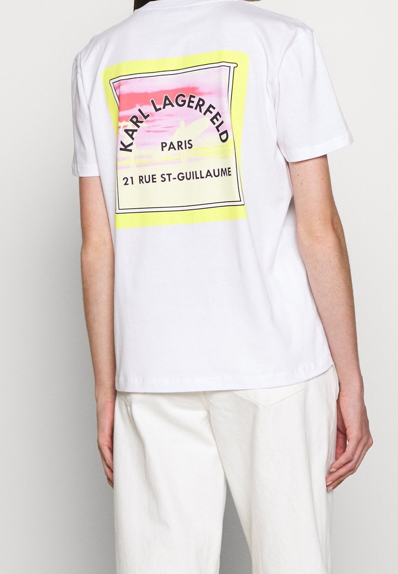 Camiseta de algodón blanca con un gráfico colorido en la parte posterior que presenta "KARL LAGERFELD", París y una dirección en un marco rectangular.