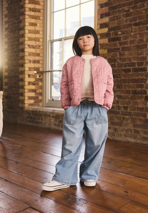 Nuori tyttö seisoo sisällä käsineen leveälahkeisten farkkujen taskuissa, hänellä on päällä vaaleanpunainen tikattu takki ja valkoiset lenkkitossut puulattialla.