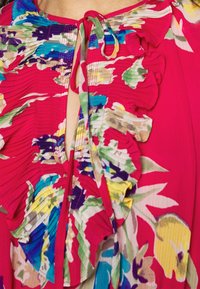 Světlé růžové tričko s květinovými vzory, skládací textura, volánky kolem výstřihu a zavazování na přední straně. Obsahuje různé barvy.