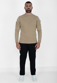 Maglione beige a maglia con polsini e orlo a costine, abbinato a pantaloni neri e sneakers blu chiaro, in piedi su uno sfondo bianco.