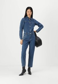 Macacão de ganga azul com fecho de botão à frente, colarinho e bolsos à frente. Combinado com botas de tornozelo pretas e uma mala preta.
