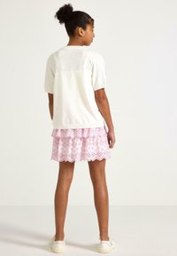 Pullover bianco leggero con maniche corte abbinato a una gonna rosa con volant caratterizzata da dettagli in occhiello e orlo scallop, indossato con scarpe bianche.
