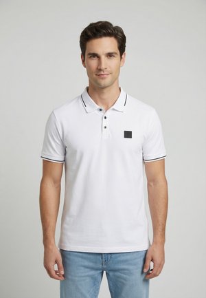 Uomo con polo bianca a maniche corte con bordi neri e jeans azzurri, in piedi davanti a uno sfondo grigio chiaro uniforme.