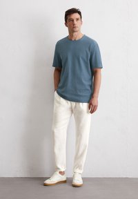 Lichtblauw t-shirt met korte mouwen en een ronde hals, gecombineerd met crèmekleurige broek en witte sneakers met tan kleurige zolen, op een neutrale achtergrond.