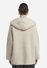 Beige stickad huva tröja med avslappnad passform, texturerad tyg, nedfällda axlar och öppna sidor. Har en stor, rundad huva.