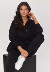 Czarna, oversize bluza z kołnierzykiem i detalami zapinanymi na guziki, zestawiona z czarnymi spodniami i białymi trampkami. Gładki materiał i luźny krój.