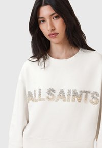 Felpa color crema con scollatura rotonda e vestibilità oversize, caratterizzata da un logo "ALLSAINTS" in rilievo e perline in toni di argento sfumati.