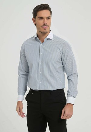 Homme portant une chemise de ville à rayures bleu clair avec col et poignets blancs, associée à un pantalon noir, debout devant un fond uni.