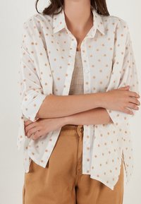 Personne portant une chemise blanche à boutons avec de petits motifs d'étoiles marron et des manches retroussées par-dessus un haut côtelé beige et un pantalon marron clair.