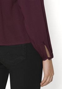 Maglietta bordeaux a maniche lunghe con una fessura sul polsino, abbinata a jeans neri attillati con una cucitura decorativa sulla tasca posteriore.