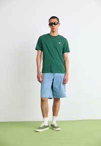 Grön bomullströja med en bröstlogotyp, matchad med ljusblå denimshorts och gröna sneakers. Modellen bär svarta solglasögon och vita strumpor.