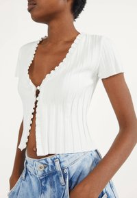 Weißes, geripptes Kurzarmoberteil mit einem rüschengestalteten Ausschnitt und vorderen Knöpfen. Das Design zeichnet sich durch eine figurbetonte Form und einen strukturierten Stoff aus.