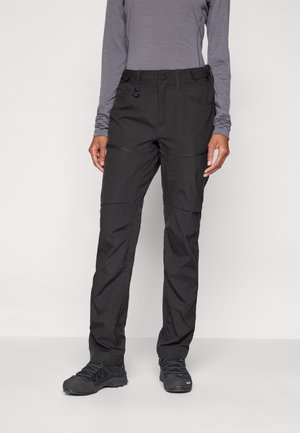 Person trägt schwarze Outdoor-Hose, graues Langarmshirt und schwarze Wanderschuhe und steht vor einem einfarbigen Hintergrund.