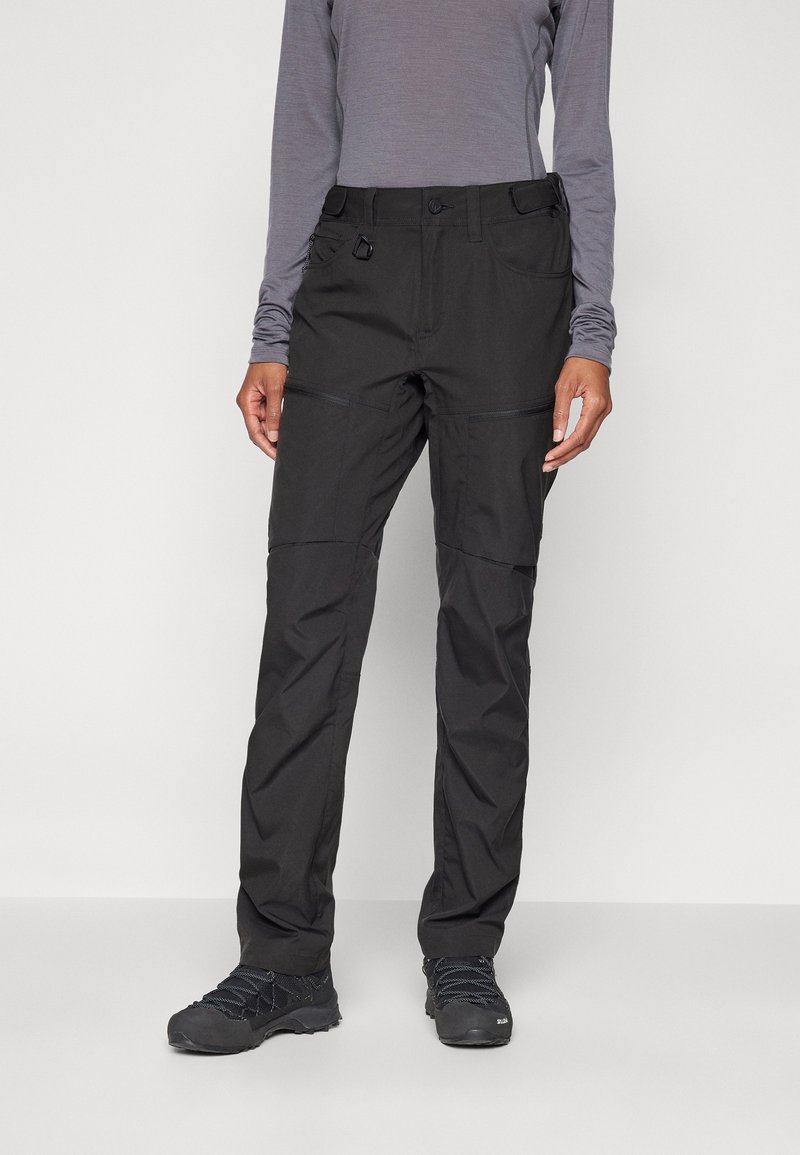 Personne portant un pantalon de randonnée noir, un haut gris à manches longues et des chaussures de randonnée noires, debout devant un fond uni.
