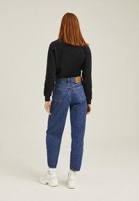 Högmidjade blå jeans i denim med avslappnad passform, med en läderlogotyp på baksidan och vita sneakers.