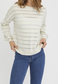 Frau in einem weißen Strickpullover mit metallischen horizontalen Streifen und blauen Jeans, die die untere Kante des Pullovers mit den Händen hält.