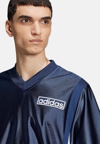 Marinblå Adidas-tröja med V-hals, tillverkad i glänsande tyg. Har vitt logo på bröstet och texturerade sidopaneler.