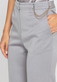 Jasnoszare dopasowane spodnie o gładkiej fakturze, z bocznymi kieszeniami i metalicznym łańcuchem na pasku dla dodatkowego akcentu.