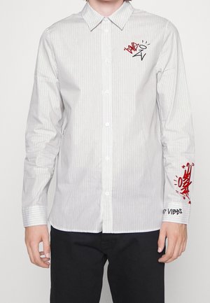 Biała koszula w cienkie paski z długim rękawem, z czerwono-czarnym napisem w stylu graffiti na piersi i lewym mankiecie, noszona z czarnymi spodniami.