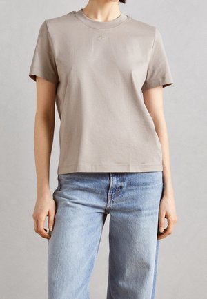 Persoon die een effen beige korte mouwen T-shirt en lichtblauwe high-waisted spijkerbroek draagt tegen een neutrale achtergrond.