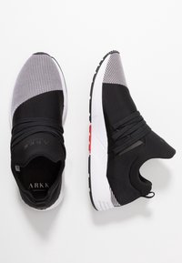 Mustat ja valkoiset liukuvoitelukengät, joissa on elastiset nauhat, neulottu tekstuurinen varvasosa ja valkoiset pohjat, joissa on pieniä punaisia yksityiskohtia, esitelty ylhäältä ja sivulta.