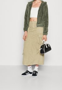 Zielona, puszysta kurtka zapinana na zamek, biała crop top, beżowa długa spódnica z kieszeniami, czarne sneakersy z białymi paskami oraz mała czarna torebka.