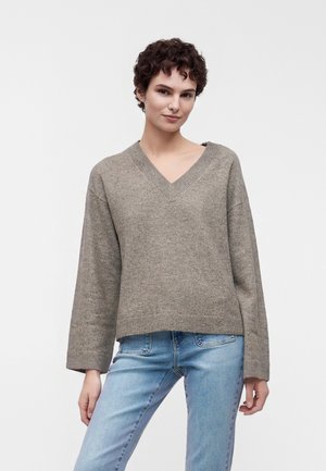 Ung person med kort krøllet hår iført en løs grå V-hals trøje og lyseblå jeans mod en ensfarvet baggrund.