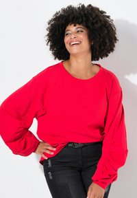 Roter, übergroßer Sweatshirt mit Raglanärmeln, ausgestattet mit gerafften Details an den Bündchen. Kombiniert mit dunklen, ausgebleichten Jeans und einem schwarzen Gürtel.