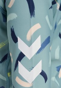 Ljusblått tyg med abstrakta penseldragmönster i grönt, marinblått och rosa, med framträdande vita chevronmarkeringar.