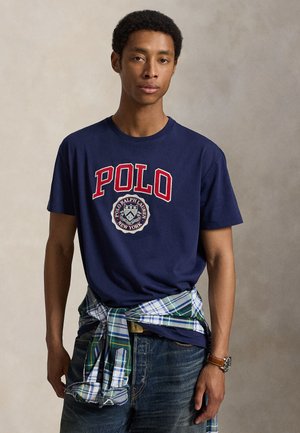 Młody mężczyzna w granatowej koszulce Polo Ralph Lauren i dżinsach, z zielono-niebieską koszulą w kratę zawiązaną wokół talii.