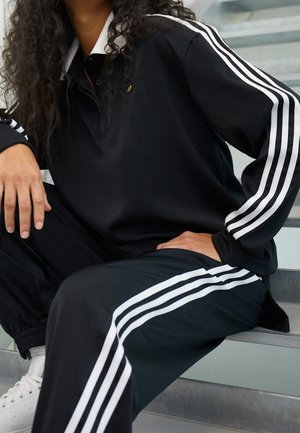 Personne assise sur des escaliers portant un survêtement noir avec des bandes blanches sur les manches et le pantalon, assorti à des baskets blanches.