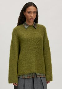 Grüner, flauschiger übergroßer Pullover mit rundem Halsausschnitt und langen Ärmeln, über einem karierten Hemd mit Kragen getragen, kombiniert mit einem grauen Faltenrock.