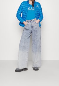 Modrá péřová bunda, světle modré tričko s logem, světle šedé džíny s širokými nohavicemi a černé lesklé boty.