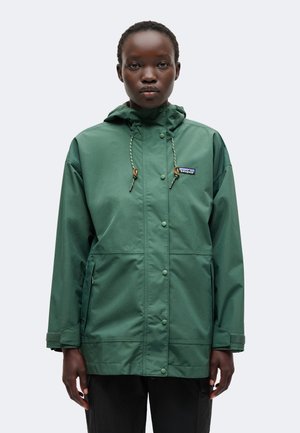 Adulte portant une veste de pluie verte à capuche avec fermeture à boutons et zip, poignets réglables et logo Patagonia sur la poitrine.