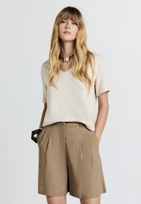 Blouse beige à manches courtes avec un décolleté en V, associée à un short brun ajusté avec des plis devant et des poches latérales. Accentuée par un collier imposant.