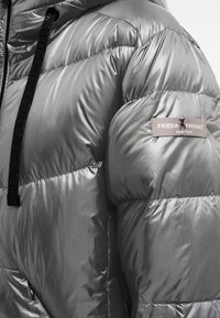 Silberne Pufferjacke mit glatter, gesteppten Textur. Verfügt über schwarze Kordelzüge und ein Logopatch am Ärmel.