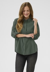 Dark green button-up shirt with a collar, long sleeves rolled to the elbows, and a relaxed fit. Cotton-like texture and rounded hem.
