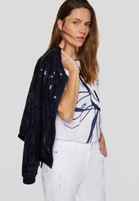 Black sequined cardigan worn over a white t-shirt with a blue abstract design. White distressed jeans complete the outfit.