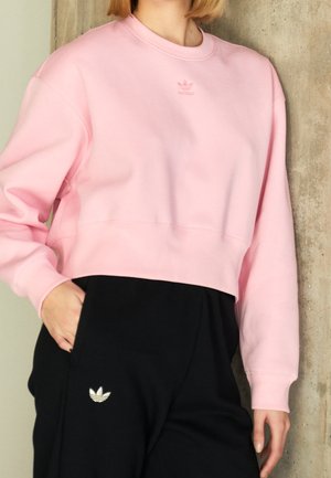 Osoba nosząca jasnoróżową krótką bluzę i czarne spodnie, oba z małymi logo Adidas trefoil, stojąca przy teksturowanej ścianie.
