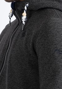 Mörkgrå hoodie gjord av texturerad tyg, med dragkedjestängning, snoddar med träpärlor och logodetaljer på framsidan.