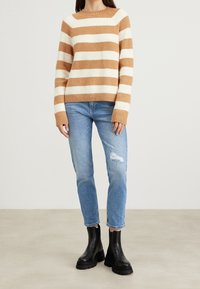 Dzianinowy sweter w szerokie paski w kolorach beżowym i kremowym, z okrągłym dekoltem i ściągaczami na mankietach, zestawiony z przetartymi jasnoniebieskimi dżinsami i czarnymi botkami za kostkę.
