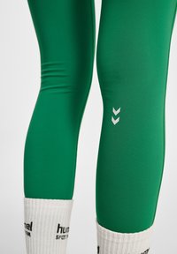 Zielone legginsy sportowe o gładkiej fakturze i dopasowanym kroju, z białym logo oraz dwoma skierowanymi w dół chevronami na udzie.